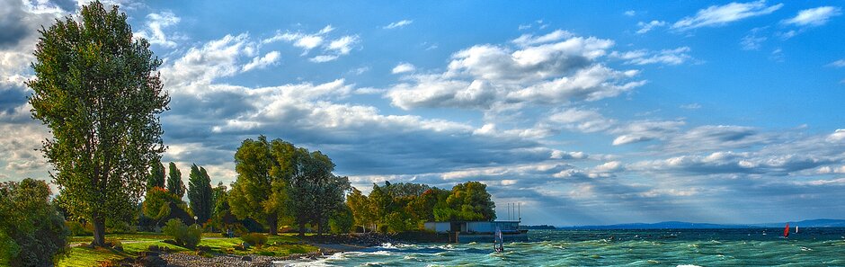 Am Bodenseeufer an einem sonnigen, leicht bewölkten Tag. Die Wellen rollen wild über den Kiesstrand, nah an die Wiese, auf welcher Bäume und Büsche wachsen.