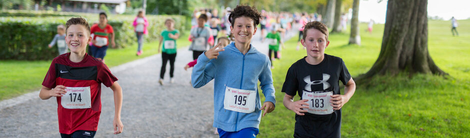 Drei Kinder, die an einem Lauf teilnehmen und joggend in die Kamera lächeln.